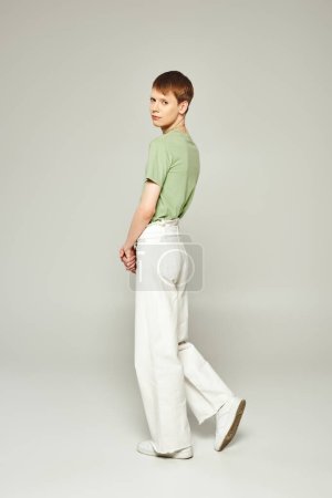 in voller Länge junger queerer Mensch mit Lipgloss, der in grünem T-Shirt und weißen Jeans steht, während er auf grauem Hintergrund in die Kamera schaut