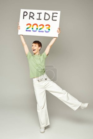 longitud completa de persona queer alegre con brillo de labios y boca abierta de pie en vaqueros blancos y camiseta verde mientras sostiene el orgullo 2023 cartel sobre fondo gris
