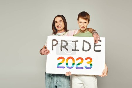 Photo for Cheerful lgbt friends holding pride 2023 placard and looking at camera while celebrating lgbtq community holiday in June on grey background in studio - Royalty Free Image
