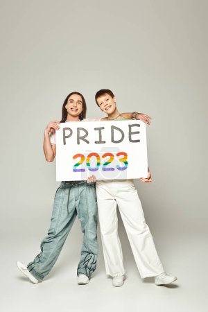 Photo for Happy lgbt friends holding pride 2023 placard and looking at camera while celebrating lgbtq community holiday in June on grey background in studio - Royalty Free Image