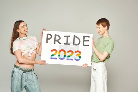 Foto de Alegre gay los hombres celebración orgullo 2023 cartel mientras celebrando lgbtq comunidad día de fiesta en junio y de pie juntos en gris fondo en estudio - Imagen libre de derechos