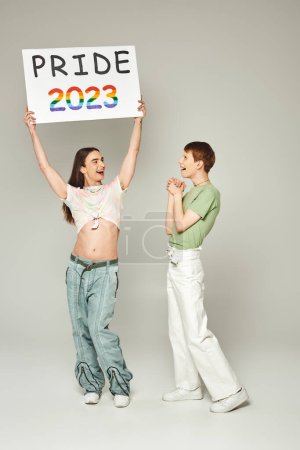 happy gay man with bare belly holding pride 2023 placard while standing next to amazed queer friend and celebrating lgbtq community holiday in June on grey background in studio 