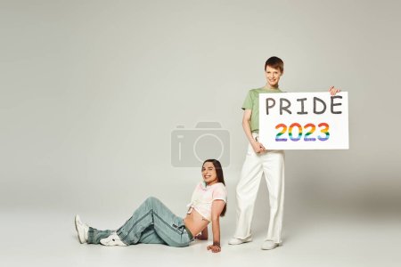 happy gay man holding pride 2023 placard while standing next to smiling queer friend with bare belly and celebrating lgbtq community holiday in June on grey background in studio 