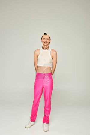 full length of happy and tattooed gay man in baseball cap, crop top and pink pants smiling while posing on grey background, pride day concept 