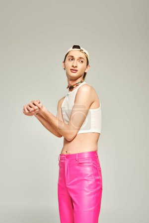 tattooed young gay man in baseball cap, crop top, bare belly and pink pants posing with clenched hands and looking away on grey background, pride day concept 