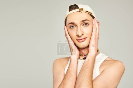 retrato de hombre gay joven tatuado con ojos azules posando en gorra de béisbol y cuentas de colores y mirando a la cámara en el fondo gris, concepto del día del orgullo 