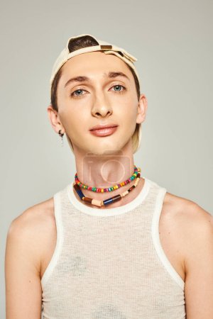 portrait of young gay man with blue eyes eyes posing in baseball cap, white tank top and colorful beads on grey background, pride day concept 