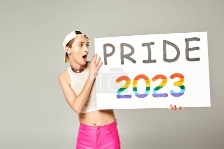tattooed and shocked gay man in baseball cap, crop top, and pink pants holding pride 2023 placard while standing with opened mouth on grey background 