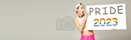 tattooed and shocked lgbt community man in baseball cap, crop top, and pink pants holding pride 2023 placard while standing with opened mouth on grey background, banner  