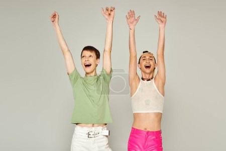 happy lgbt friends in colorful clothes standing with raised hands while celebrating lgbtq community holiday in June on grey background in studio, pride month concept 