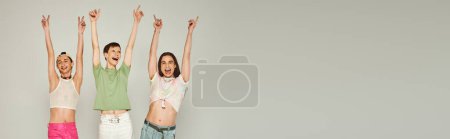 happy and young lgbt community friends in colorful clothes smiling while looking at camera, raising hands and celebrating pride month together on grey background in studio, banner 