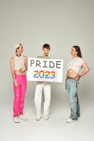 happy gay men in colorful clothes standing and looking at queer friend holding pride 2023 placard while celebrating lgbt community holiday in June, grey background, studio 