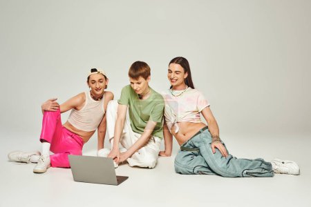 Diverse positive und junge lgbt-Freunde mit Tätowierungen sitzen in bunten Klamotten zusammen und benutzen Laptop im Studio auf grauem Hintergrund während des Monats Stolz 