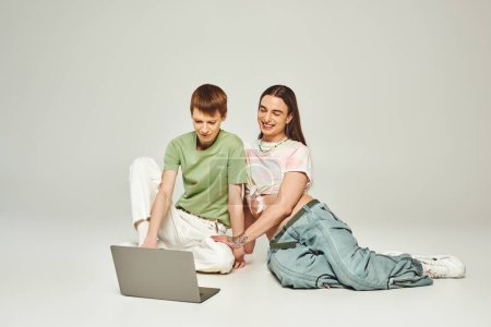 positiver und junger schwuler Mann in bunten Klamotten sitzt neben tätowiertem Freund und benutzt Laptop zusammen im Studio auf grauem Hintergrund während des Monats des Stolzes 