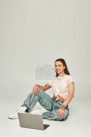 positive and young gay man with long hair and trendy clothes sitting and looking at modern laptop in studio on grey background during pride month 