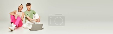 junger schwuler Mann in bunten Klamotten sitzt neben Freund mit Baseballmütze und schaut Laptop zusammen im Studio auf grauem Hintergrund während des Monats Stolz, Banner 