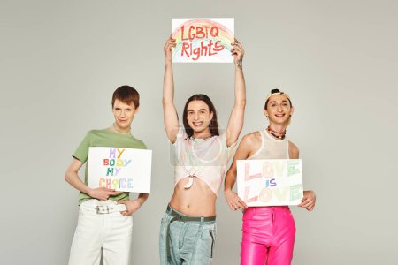 happy lgbt friends holding placards with my body my choice, lgbtq rights and love is love lettering while standing together on pride month, grey background 
