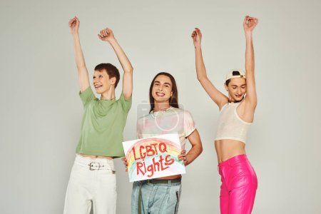 Foto de Amigos lgbt feliz en ropa colorida bailando con las manos levantadas junto a activista sosteniendo cartel con letras derechos lgbtq y sonriendo en el mes de orgullo, fondo gris - Imagen libre de derechos