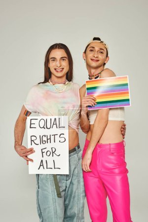 happy and tattooed lgbt friends holding rainbow flag picture and placard with equal rights for all lettering while standing together on pride day, grey background 