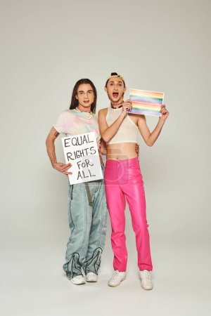 Photo for Emotional and tattooed lgbt people holding rainbow flag picture and placard with equal rights for all lettering while standing together on pride day, grey background - Royalty Free Image