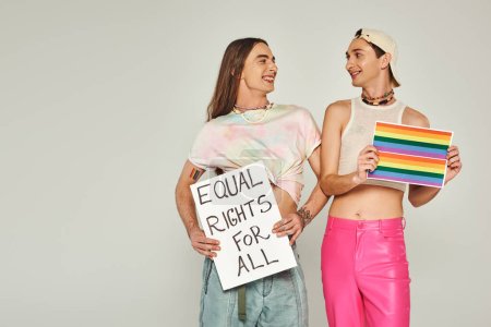 Photo for Positive and tattooed lgbt people holding rainbow flag picture and placard with equal rights for all lettering while looking at each other on pride day, grey background - Royalty Free Image