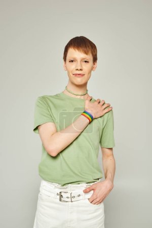 Foto de Retrato de joven queer con brillo de labios de pie en camiseta verde y mostrando la mano con lgbt brazalete de la bandera mientras mira a la cámara durante el mes de orgullo sobre fondo gris - Imagen libre de derechos