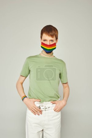 Foto de Retrato de una joven queer de pie en camiseta verde y máscara de bandera lgbt mientras mira a la cámara y posando durante el mes de orgullo sobre fondo gris - Imagen libre de derechos