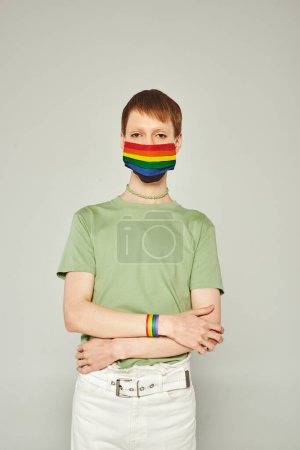 retrato de un joven gay de pie en camiseta verde y máscara de bandera lgbt mientras mira a la cámara y posando durante el mes de orgullo sobre fondo gris
