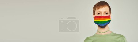 Foto de Retrato de una joven queer de pie en camiseta verde y máscara de la bandera del arco iris lgbt mientras mira a la cámara durante el día del orgullo sobre fondo gris, pancarta - Imagen libre de derechos