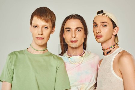 portrait of three lgbtq people with colorful beads looking at camera while standing together on grey background in studio, celebration of pride month concept  
