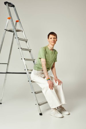 Photo for Full length of happy queer person with shiny lip gloss posing in green t-shirt and white denim jeans while sitting on ladder during pride month on grey background - Royalty Free Image