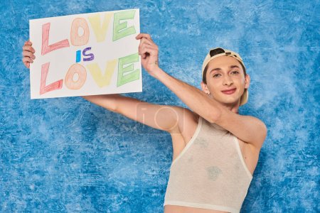 cheerful young gay activist in baseball cap and white tank top smiling while holding placard with love is love words during pride month on mottled blue background 