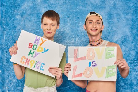 positive schwule Aktivisten lächeln, während sie Plakate mit Liebe halten, ist Liebe und mein Körper meine Lieblingsworte während des stolzen Monats auf fleckigem blauem Hintergrund 