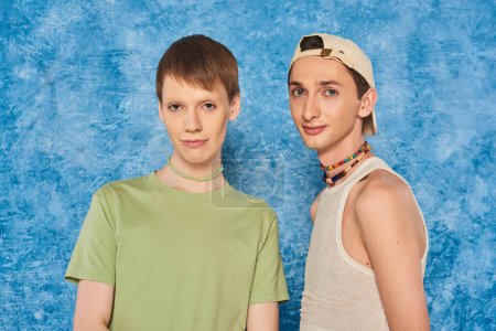 young lgbtq friends with colorful beads and casual clothes and looking at camera while standing together on mottled blue background during pride month 