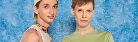 young lgbtq friends with colorful beads and casual clothes looking at camera while standing together on mottled blue background during pride month, banner 