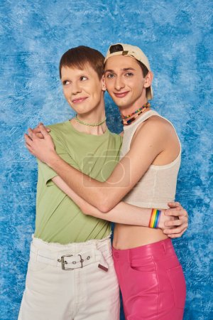 Foto de Jóvenes amigos lgbtq con cuentas de colores de pie en ropa casual y abrazándose mientras sonríen sobre fondo azul moteado durante el mes de orgullo - Imagen libre de derechos