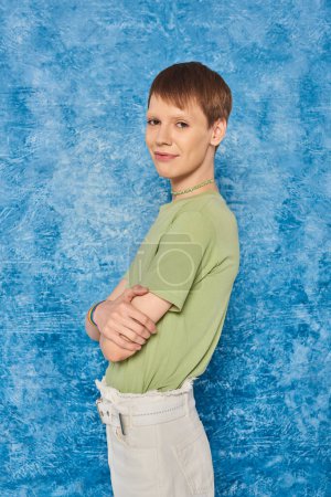 Smiling queer person in casual clothes crossing arms and smiling at camera while celebrating pride month and standing on mottled blue background