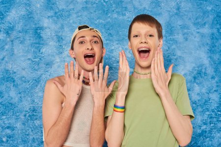 Amigos homosexuales conmocionados con ropa casual abriendo la boca y mirando a la cámara durante la celebración del mes de orgullo lgbt sobre fondo azul moteado