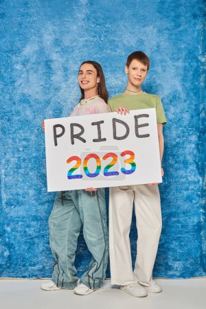 Full length of cheerful queer friends looking at camera while holding placard with pride 2023 lettering during lgbt month celebration on mottled blue background