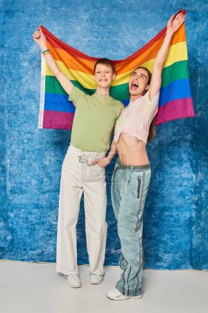 Foto de Longitud completa de la comunidad homosexual emocionada con ropa casual sosteniendo la bandera lgbt mientras celebran el mes de orgullo y de pie juntos sobre fondo azul moteado - Imagen libre de derechos