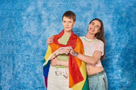 Foto de Sonriente hombre gay tatuado en ropa casual abrazando a un joven amigo con bandera lgbt y mirando a la cámara durante la celebración del mes de orgullo sobre fondo azul moteado - Imagen libre de derechos