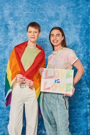 Carefree queer community with lgbt flag holding placard with love is love lettering and looking at camera while celebrating pride month on mottled blue background