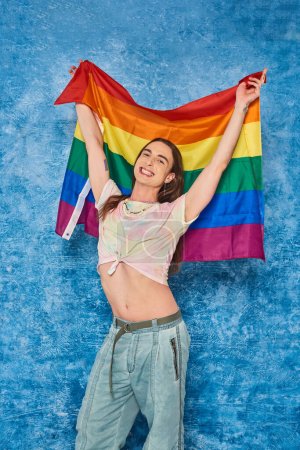 Cheerful long haired and tattooed homosexual man raising lgbt flag during pride community month celebration on textured  mottled blue background