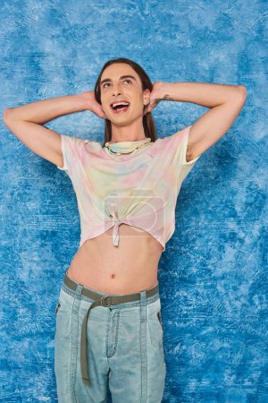 Cheerful and dreamy homosexual man in top and jeans touching hair and looking away during lgbt month celebration on mottled blue background