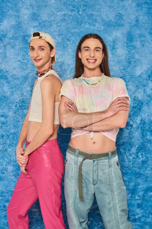 Stilvolle und junge homosexuelle Freunde posieren und lächeln gemeinsam in die Kamera während der lgbt Community Priority Monatsfeier auf texturiertem blauem Hintergrund