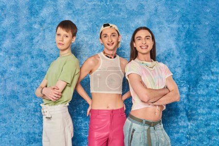 Cheerful and stylish homosexual friends looking at camera together and posing while celebrating lgbt community month on mottled blue background