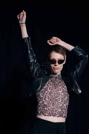 Trendy und junger homosexueller Mann in Sonnenbrille, Lederjacke und Bluse mit Animal Print posiert während der stolzen Monatsfeier isoliert auf schwarz 