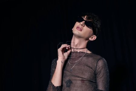 Photo for Fashionable gay man in sunglasses and sparkling top touching necklace during lgbt community and pride month party isolated on black - Royalty Free Image
