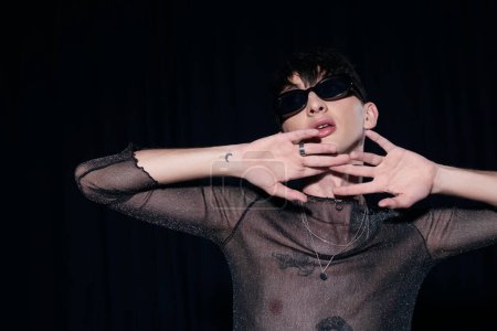 Tätowierter und modischer schwuler Mann mit Sonnenbrille und funkelndem Top, der das Gesicht während der lgbt-Monatsfeier berührt, isoliert auf schwarz 