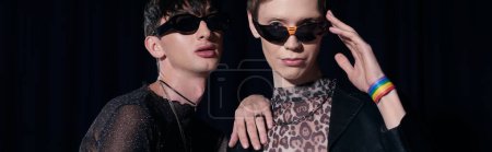 Stylish gay man in sunglasses touching shoulder of friend looking at camera during lgbt month celebration party isolated on black, banner 